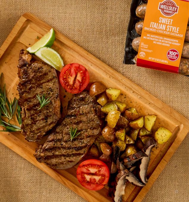 overhead photo of wellsley farms steaks on a wooden slab, with potatoes, tomatoes and italian sausage.