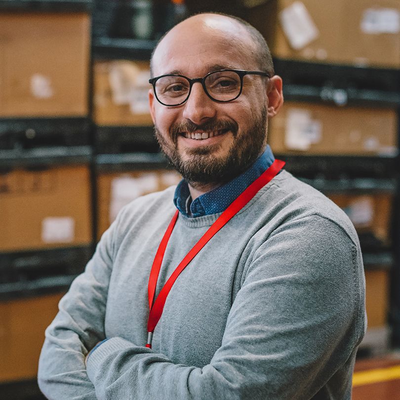 Image: Smiling man with glasses and a red lanyard standing with his arms crossed in a warehouse.