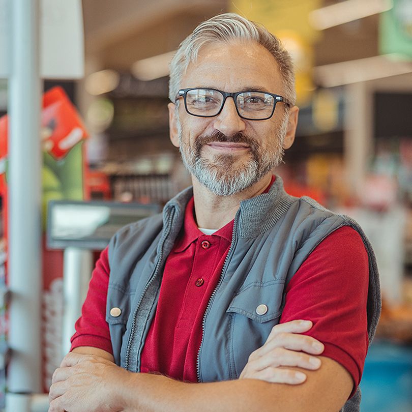 Image: Smiling man with glasses and a gray vest standing with his arms crossed in the produce section of a grocery store.