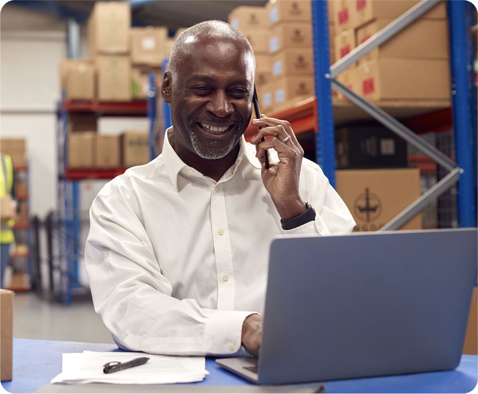 Image: Smiling man in a white shirt talking on the phone while working on a laptop in a warehouse.