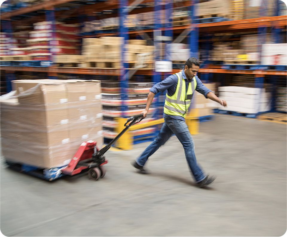 Image: Worker in a safety vest pulling a pallet jack loaded with boxes in a warehouse.