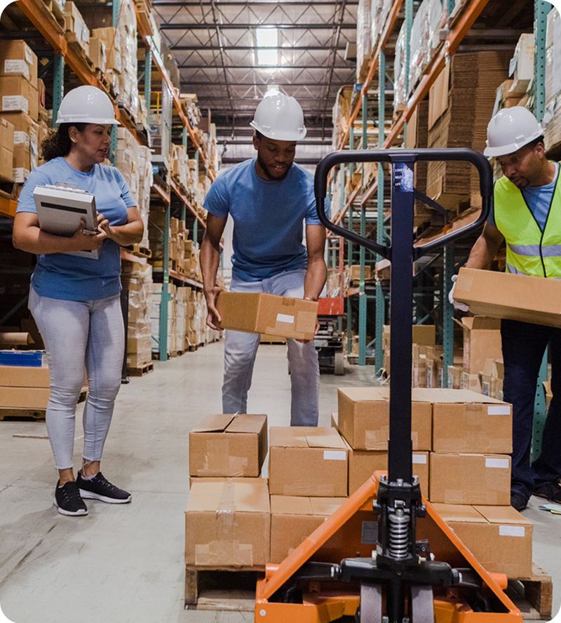 Image: Three workers in a warehouse, wearing hard hats and safety vests, handling boxes and using a pallet jack.