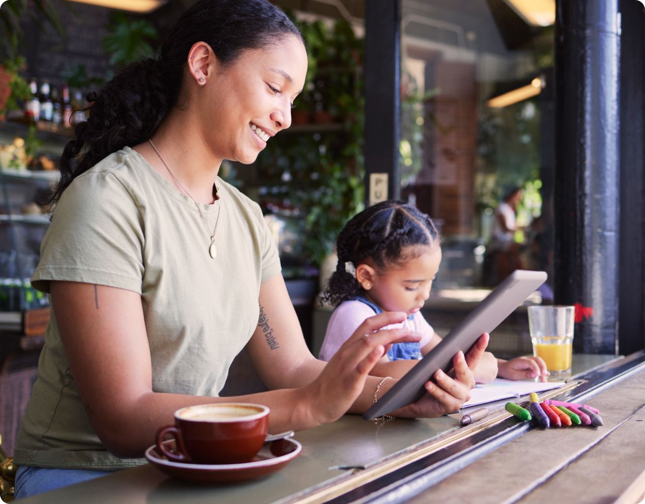 A woman looking at her tablet while her daughter sits next to her at a coffee shop.
